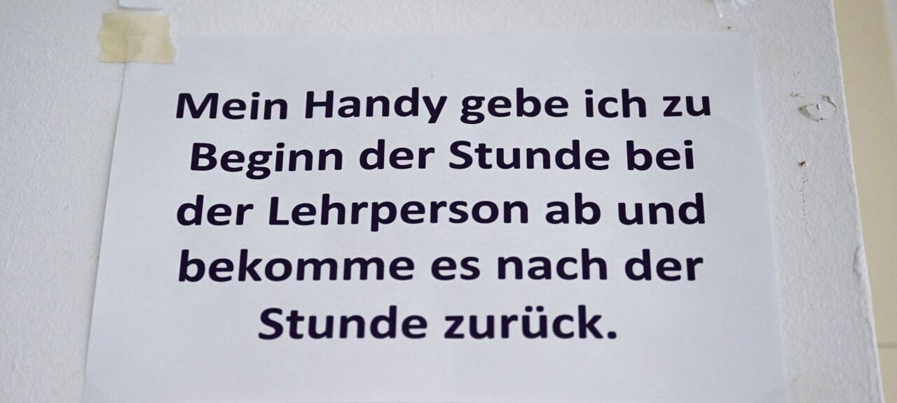 "Mein Handy gebe ich zu Beginn der Stunde bei der Lehrperson ab und bekomme es nach der Stunde zurück", steht in einem Klassenzimmer an einer Schule.
